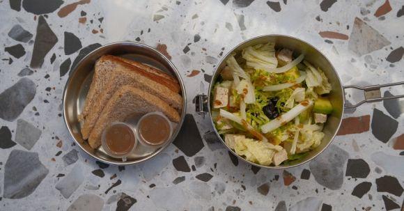 Zwei Metallboxen mit Sandwiches, Salat und zwei kleinen Bechern Dressing auf gesprenkelter Oberfläche