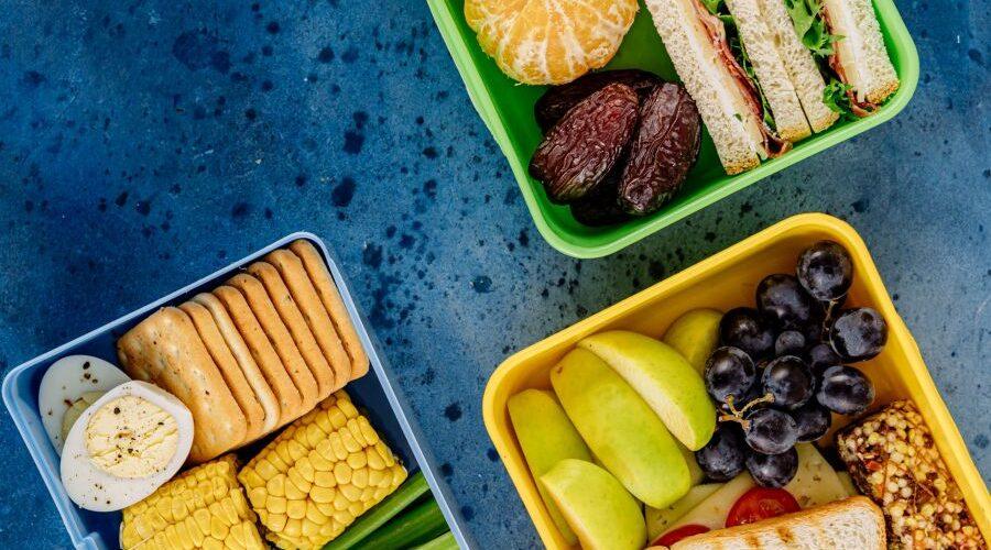 Drei geöffnete Lunchboxen mit Sandwiches, Obst, Gemüse, Crackern und Ei auf blauem Untergrund