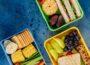 Drei geöffnete Lunchboxen mit Sandwiches, Obst, Gemüse, Crackern und Ei auf blauem Untergrund